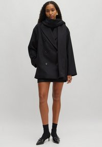 Zwarte dubbelgebreide oversized jas met een hoge kraag en grote knopen, gecombineerd met een zwarte mini-jurk, zwarte sokken en spitse hakken.