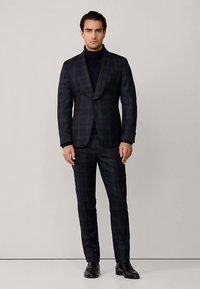 Donker ruitpak in marineblauwe en groene tinten, met een reverskraag, gecombineerd met een zwarte coltrui en zwarte schoenen. Gladde, op maat gemaakte stof.