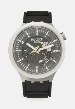 Rund sølv Swatch armbåndsur med sort transparent urskive, der viser de indvendige tandhjul, hvide timeindikatorer og en sort gummirem.