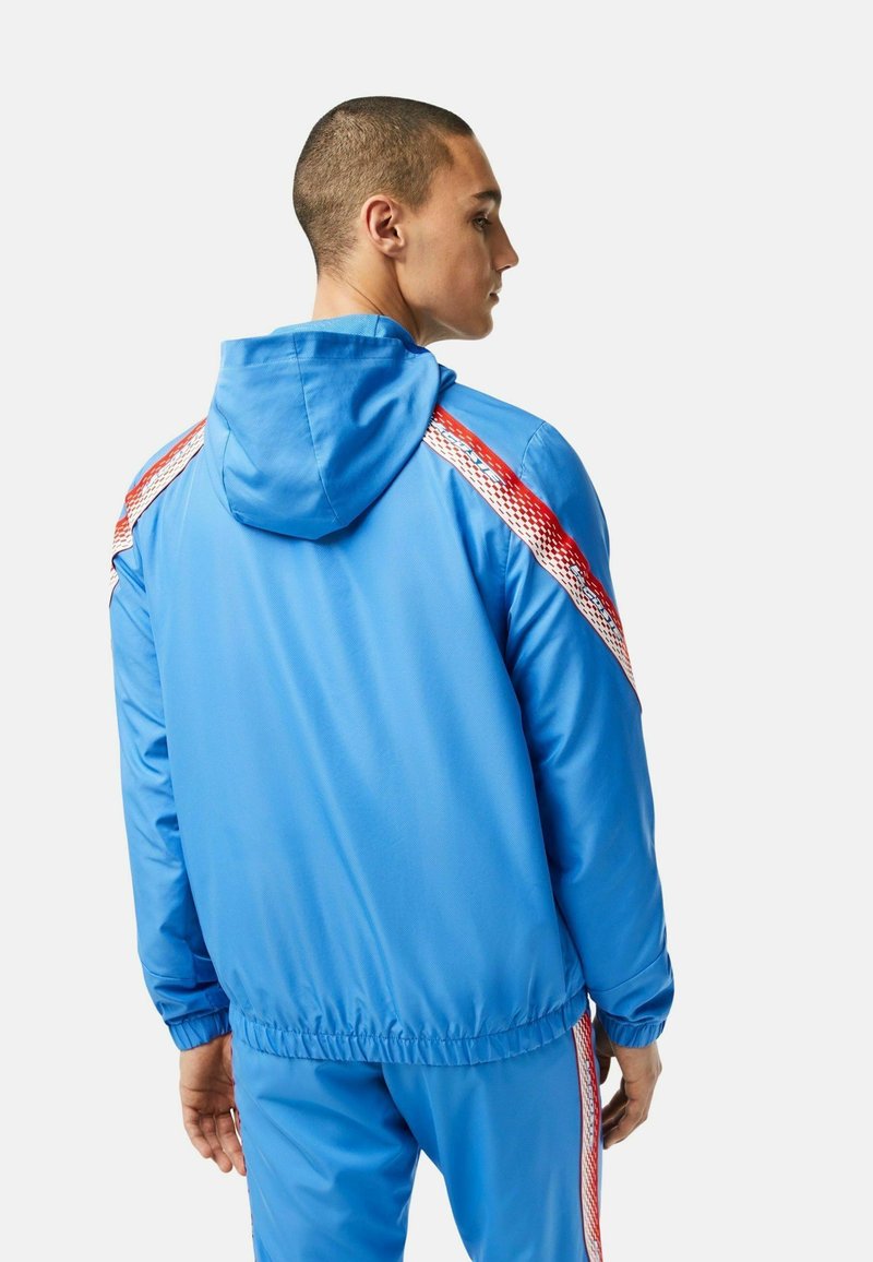 Veste de sport bleue avec capuche, présentant des rayures rouges et blanches le long des épaules. Fabriquée en matériau doux et léger avec des poignets élastiques.