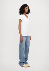 Camiseta blanca de manga corta con escote en V, combinada con unos pantalones vaqueros oversized de color azul claro que presentan un patrón de logotipos, y zapatillas beige.