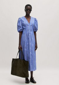 Blauw-wit gestreepte midi-jurk met pofmouwen, knoopsluiting en een diepe V-hals. Model houdt een donkergroene suède handtas vast.
