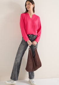 Blouse rose vif à manches longues avec col en V, associée à un jean gris délavé à jambes larges et un sac à main en daim marron, avec des accessoires métalliques.