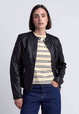 Vrouw met kort donker haar die een zwarte leren jas draagt over een crèmekleurig en zwart gestreept shirt en blauwe spijkerbroek, staand met één hand in de zak.