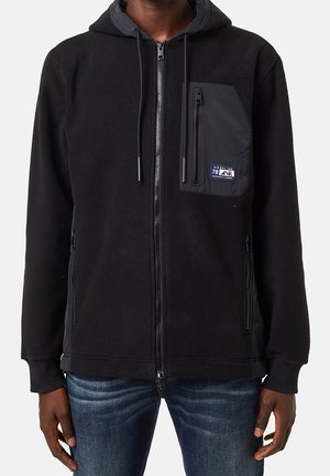 Sweat à capuche polaire noir zippé avec poche avant zippée ornée d'un petit patch logo Diesel, porté avec un jean bleu.