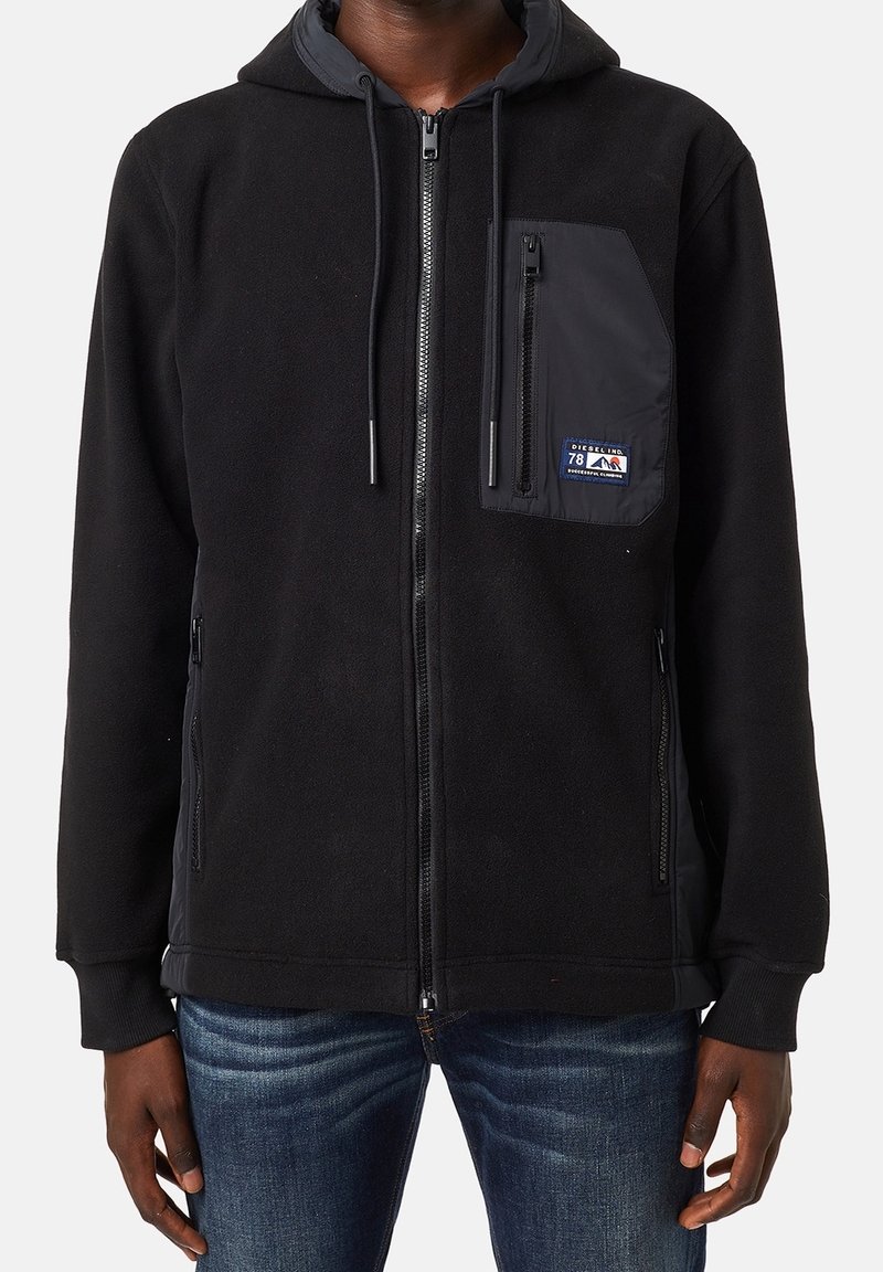 Sweat à capuche polaire noir zippé avec poche avant zippée ornée d'un petit patch logo Diesel, porté avec un jean bleu.