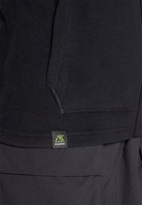 Indumento in fleece nero con texture morbida, caratterizzato da un'etichetta Polartec visibile. Design semplice, che sottolinea calore e versatilità.