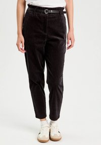 Pantalons en velours côtelé noirs avec une taille ajustée, des jambes fuselées et une ceinture noire. Présente des côtes verticales et des poches avant. Associés à des baskets blanches.