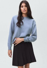 Woman with long dark hair wearing a light blue sweater and black pleated skirt, standing against a plain light gray background.