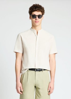 Junger Mann mit schwarzen runden Sonnenbrillen, beige Kurzarmhemd mit Mandarinkragen und hellen Khakihosen mit schwarzem Gürtel.