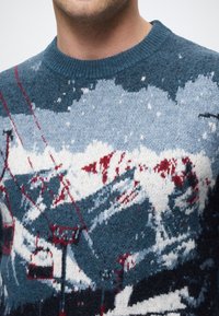 Adīts džemperis ar ainavas dizainu, kurā ir zilas, baltas un sarkanas krāsas, attēlojot kalnus un slēpošanas pacēlājus, ar apaļu kakla izgriezumu.