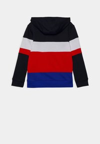 Mikina s kapucí v černé, červené, bílé a modré barvě se pruhovaným vzorem. Obsahuje dlouhé rukávy a přední klokaní kapsu.