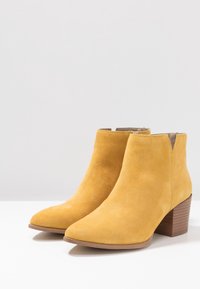 Botas de tobillo de ante amarillo con punta afilada, cortes laterales y un tacón de madera apilada. Textura suave con un acabado liso.