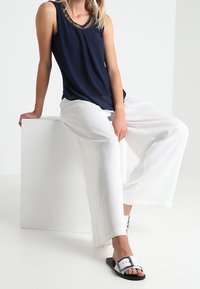 Haut sans manches bleu marine avec un col en perles, associé à un pantalon blanc à jambes larges et des sandales noires avec une boucle en argent.