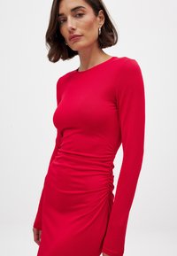 Red long-sleeve dress made of smooth fabric, featuring a fitted silhouette with side ruching details and a round neckline.