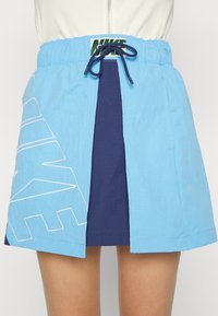 Jupe Nike bleu clair avec panneau avant bleu marine, taille à cordon, et grand logo blanc Nike sur le côté gauche, portée avec un haut blanc.