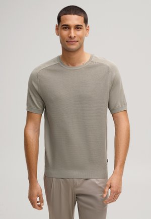 Uomo che indossa una maglia a maniche corte di maglia strutturata grigio chiaro e pantaloni grigio chiaro, in piedi davanti a uno sfondo neutro semplice.