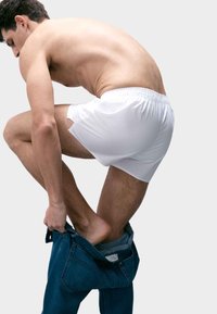 Białe bawełniane bokserki noszone przez mężczyznę, który zakłada niebieskie dżinsy. Tło jest jednolite szare.