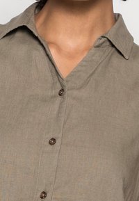 Camicia a bottoni in lino verde oliva con colletto a punta, caratterizzata da bottoni marroni e un motivo testurizzato sottile.