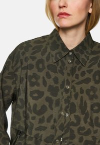 Button-up shirt in olive green with a dark leopard print pattern. Features a collar and metallic buttons, with a texture resembling lightweight fabric.