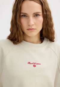 Sudadera beige con texto bordado en rojo "Plantkisses" y diseño de labios. Cuenta con un ajuste relajado y mangas raglán con una superficie texturizada.