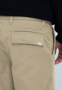 Beežid puuvillased püksid tagatasku ja väikese 'J SOLID' märgiga. Kangas on siledapinnaline ja hoolikalt istuv, vööaasadega.