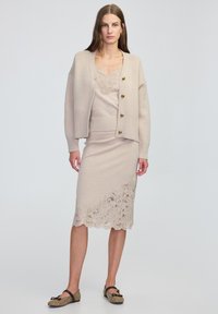 Cardigan lavorato beige con bottoni dorati, sovrapposto a un top e una gonna coordinati con dettagli in pizzo lungo l'orlo. Ballerine nere con fantasia.