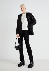 Črn usnjen blazer preko bele srajce, črne hlače in masivne črne čevlje na visokih petah, drži majhno črno torbico z teksturiranim zaključkom.