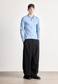 Šviesiai mėlynas ilgomis rankovėmis polo marškinėlis, derinamas su juodomis plačiomis kelnėmis ir juodais batais. Modelis taip pat dėvi juodą kepurę su „BOSS“ logotipu.