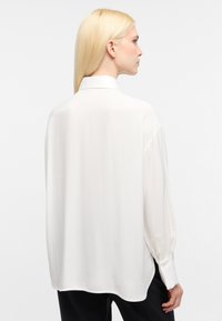 Blusa bianca a maniche lunghe realizzata in tessuto morbido. Presenta un colletto classico e polsini con bottoni. Vestibilità rilassata con un orlo leggermente curvo.
