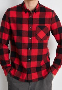 Camisa de franela a cuadros roja y negra con cuello abotonado, mangas largas y bolsillo en el pecho. Presenta botones negros y una textura suave.
