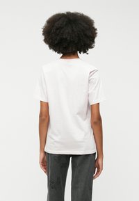 Gaiši rozā kokvilnas t-krekls ar apaļu kakla izgriezumu un īsām piedurknēm, redzams no aizmugures. Pārī ar pelēkajām biksēm ar izšūtiem rakstiem.