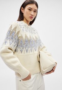 Mulher jovem a usar um suéter creme com padrão geométrico azul, segurando uma carteira triangular bege, com uma mão no bolso, contra um fundo simples.