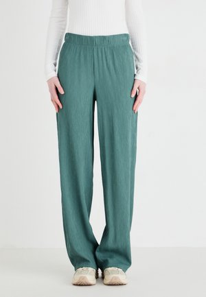 Persoon die losse tealbroeken met elastische tailleband, een witte top met lange mouwen en beige sneakers draagt, staat tegen een effen lichte achtergrond.
