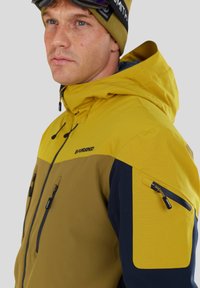 Giacca da sci gialla e verde oliva, con tasche con zip, cappuccio regolabile e dettagli a contrasto in blu navy sulle maniche.