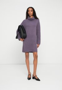 Frau in lila Rollkragenkleid mit schwarzer Umhängetasche, trägt schwarze High-Heel-Mary-Jane-Schuhe und steht vor weißem Hintergrund.