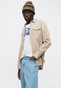 Mladi moški nosi bež srajco na gumb over belo grafično majico, svetlo modre jeans hlače in prugasto pleteno kapo v rjavih in črnih tonih.