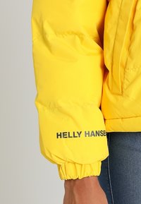 Manche de doudoune jaune Helly Hansen avec un poignet élastique portée sur un jean en denim bleu foncé.