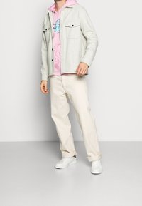 Ljusgrå knappen-up skjorta över en rosa hoodie med grafisk design, krämfärgade byxor och vita sneakers. Avslappnad outfit med lager av texturer.