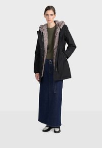 Černá parka s kapucí lemovanou kožešinou, v kombinaci s olivově zeleným žebrovaným svetr, dlouhou džínovou sukní s knoflíkovým detailem a kotníkovými botami.