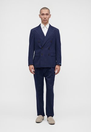 Ein Mann steht in einem marineblauen Nadelstreifen-Doppelreiheranzug mit weißem Hemd und beigen Loafers vor einem schlichten hellen Hintergrund.