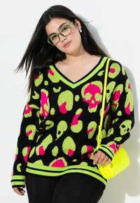 Schwarzer Pullover mit neon grünen und pinken Totenkopf- sowie Leopardenmuster; mit V-Ausschnitt und gestreiften Bündchen. Die Accessoires umfassen eine gelbe Umhängetasche.