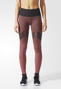 Leggings en rojo oscuro con un patrón texturizado y moteado; cuenta con paneles de malla negra y una cintura alta y acanalada. Se usan con zapatillas deportivas negras.