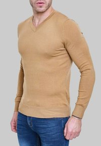 Pull en V couleur brun clair, fabriqué dans un tissu en maille lisse. Il a des manches longues et une coupe ajustée, présenté avec un jean bleu.