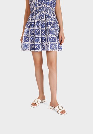 Donna che indossa un vestito corto con motivo blu e bianco e sandali bianchi senza lacci con una fibbia circolare dorata, in piedi su uno sfondo bianco.