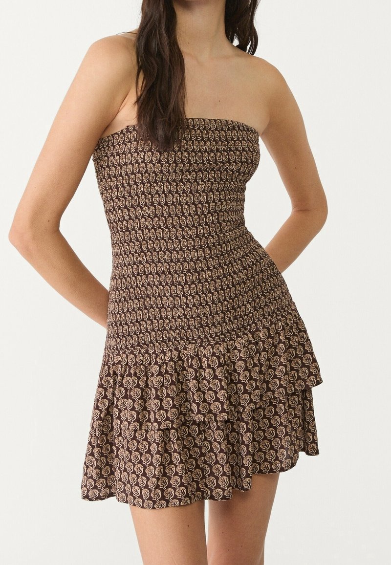 Femme portant une mini-robe bustier marron avec un motif floral beige et un ourlet volanté, debout, les bras derrière le dos.