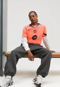 Nike Barselonos marškinėliai ryškiai oranžinės spalvos su mėlynomis detalėmis, pilkos sportinės kelnės ir sidabrinės sportinės avalynės. Marškinėliuose yra logotipas ir Spotify ženklinimas.