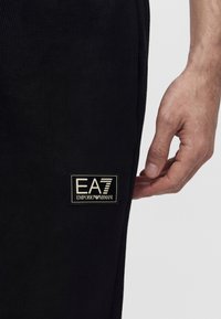Κοντινή λήψη ενός χεριού δίπλα σε μαύρο παντελόνι με ορθογώνιο έμπλαστρο λογοτύπου EA7 Emporio Armani στο μηρό.