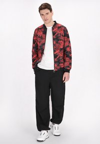 Rode en zwarte bomberjack met een palmbomenpatroon, gedragen over een wit T-shirt, gecombineerd met zwarte broek en witte sneakers.