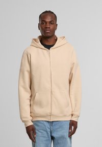 Beige zip-up hættetrøje lavet af blødt stof, med snor i hætten, lange ærmer og frontlommer. Glat tekstur, afslappet pasform.